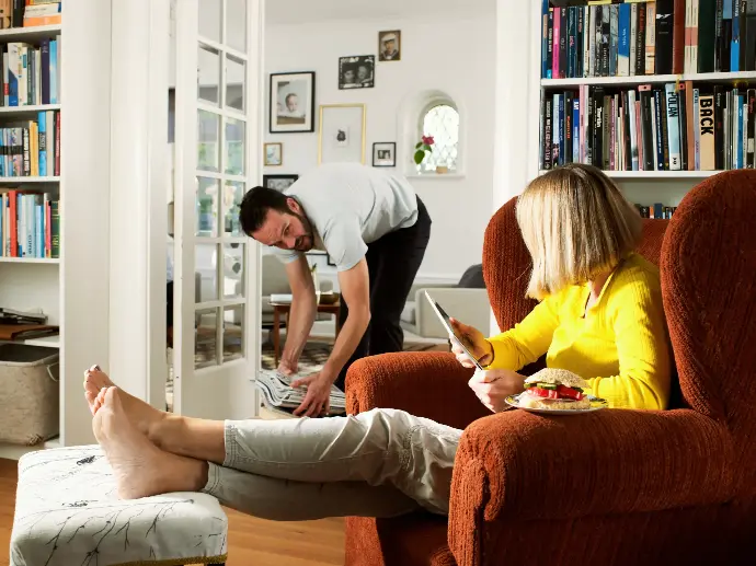 Pareja en el salón, la mujer está sentada en un sillón y el hombre recogiendo unas revistas del suelo.